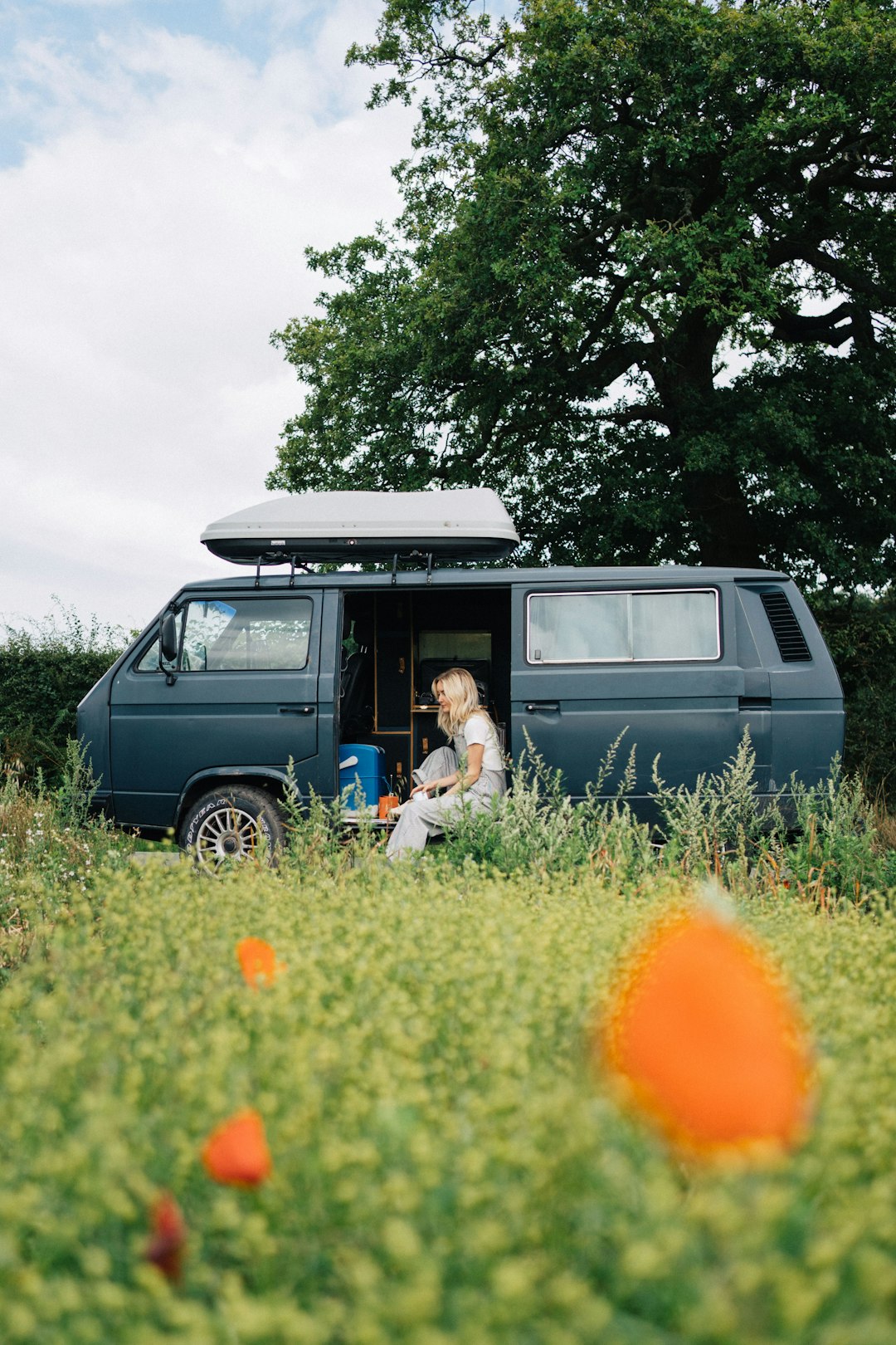 A blonde girl sitting in a camper van in a bright green field with red flowers, on a sunny day.